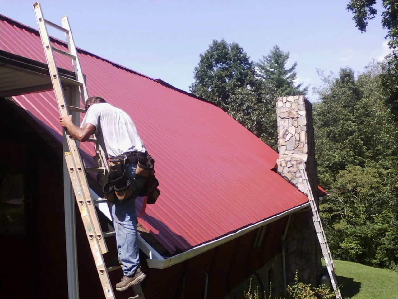 Roofer on a ladder beside a red metal roof and stone chimney — Soffit & Fascia Repair by Roofing Brothers in Chagrin Falls
